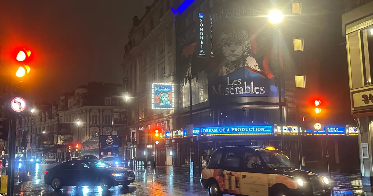 A London taxi on a wet street in Theatreland at night, the lit theatre fronts behind it